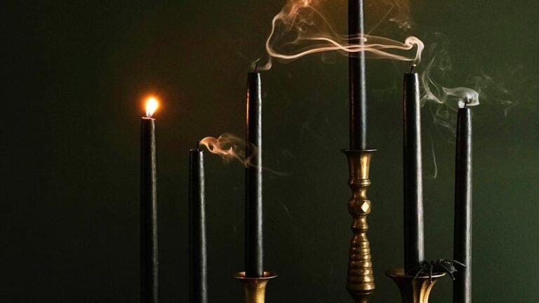 A lit black taper candle beside two smoking candles in antique brass holders against a dark green wall