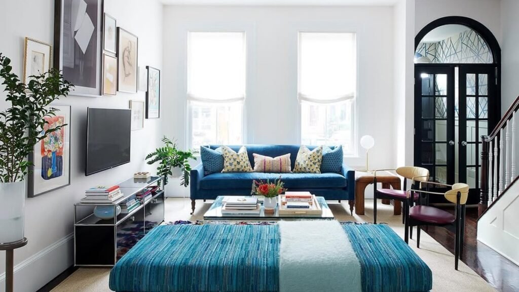 Narrow living room with a cobalt velvet sofa and matching striped ottoman, a low media console anchoring the long wall beneath a gallery of framed art