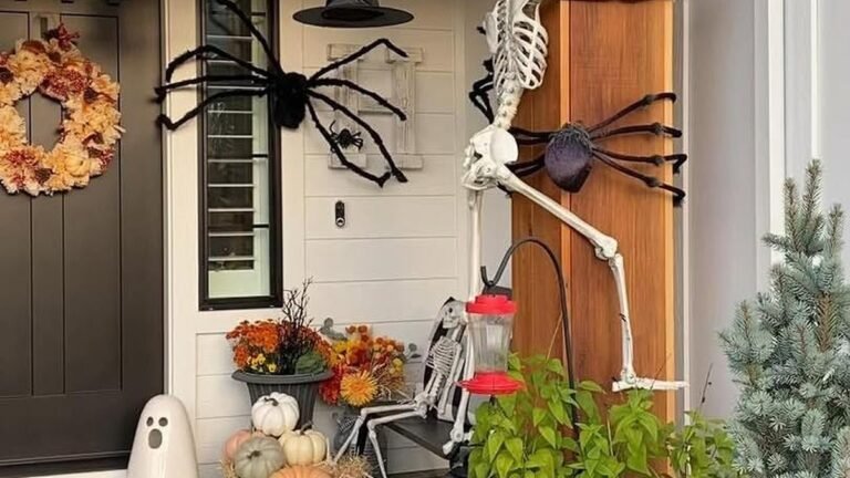 A lifelike white skeleton climbs a wooden porch post next to a large black spider, with a jack-o'-lantern, orange and green pumpkins, fresh mums, and witch hats decorating a front porch entryway