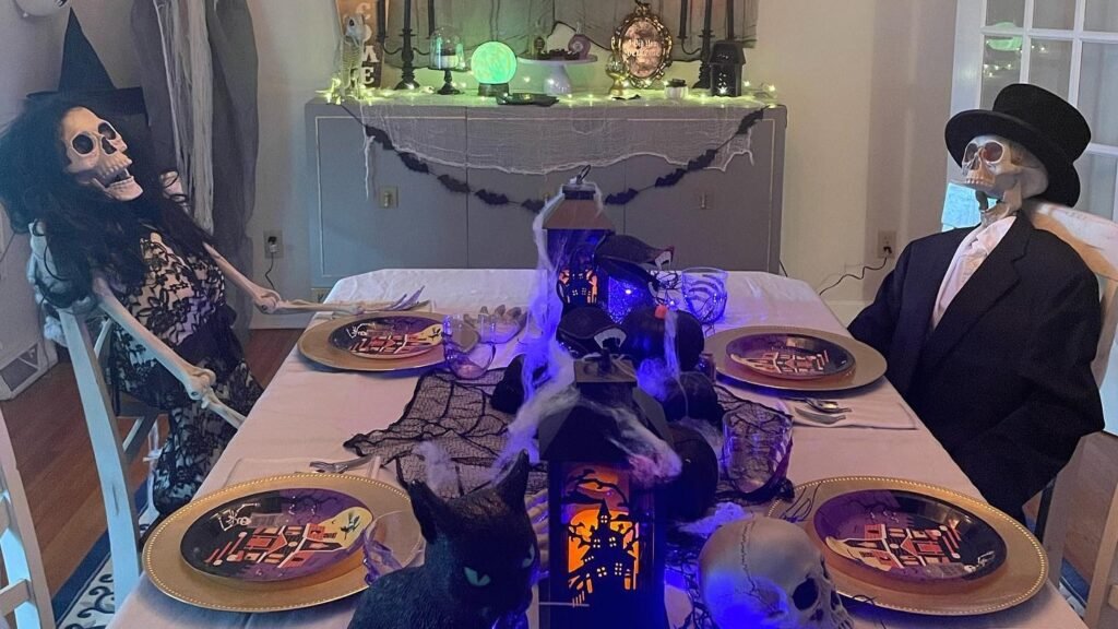 Halloween dinner table set with skeleton figures in formal attire, gold charger plates with Halloween-print dinnerware, and a glowing lantern centerpiece with cobweb styling