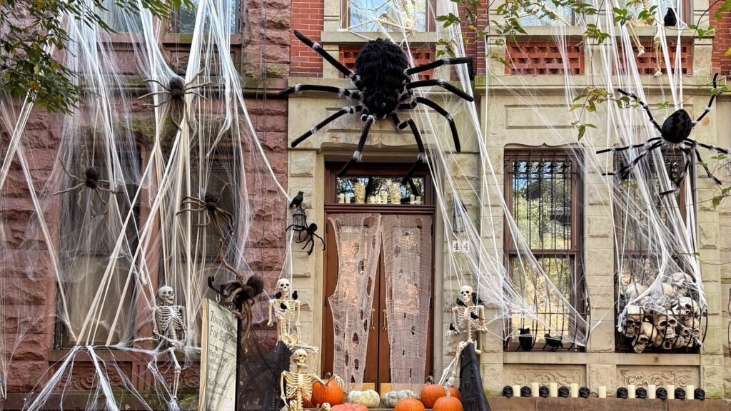 A giant black spider perches above a brownstone's lit doorway, with skeleton figures in the windows and pumpkins lining the staircase below for a dramatic Halloween entryway