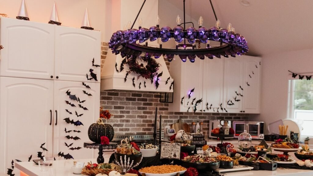 Purple skull string light chandelier hanging over a Halloween-decorated kitchen island with black bat decals on white cabinets and a full grazing table spread