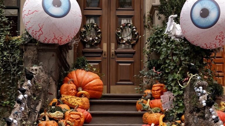 Brownstone stairway completely covered in stacked orange, cream, and green pumpkins and gourds, with two giant inflatable eyeball decorations hanging above the doorway