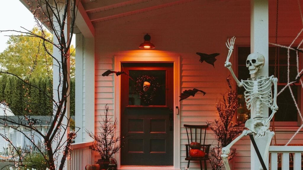 White porch glowing warm red at dusk with a skeleton climbing the railing beside stacked heirloom pumpkins in dusty rose and sage