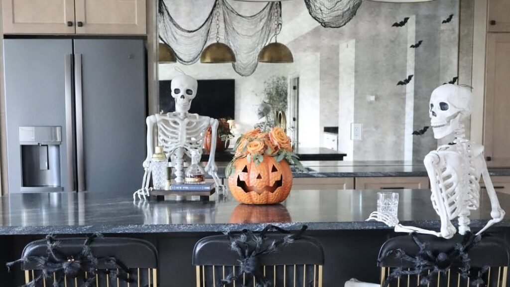 Two skeleton decorations seated at a black and gold kitchen island bar, with a jack-o'-lantern floral arrangement and black cheesecloth draped from gold pendant lights overhead