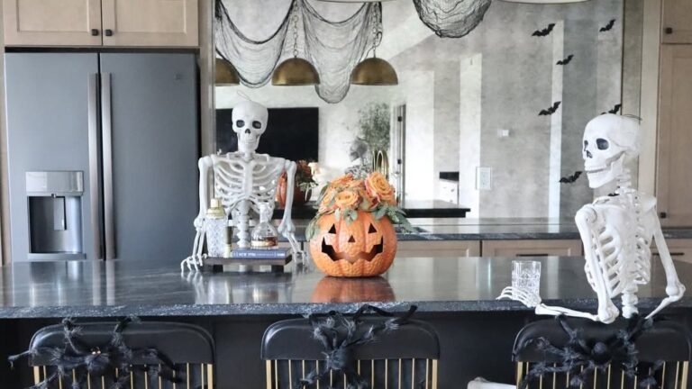 Two skeleton decorations seated at a black and gold kitchen island bar, with a jack-o'-lantern floral arrangement and black cheesecloth draped from gold pendant lights overhead