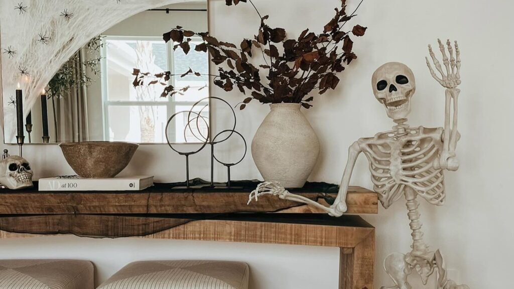 Halloween entry table decor with a life-size skeleton seated on a stool beside a wood console, cobweb-draped arched mirror, and dried branches in a ceramic vase