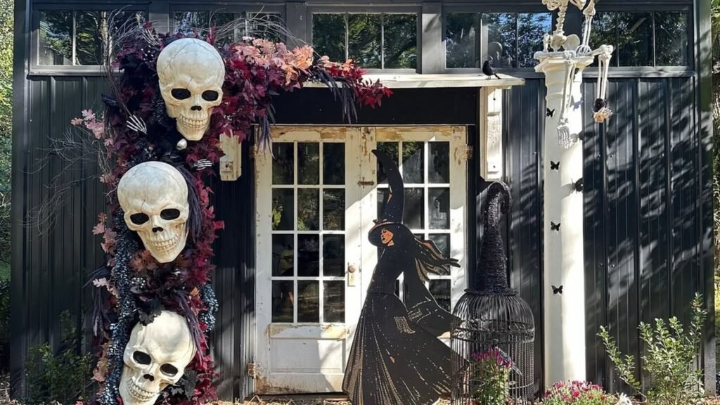 Black backyard shed decorated for Halloween with three oversized skulls, a skeleton figure on the roofline, and a dancing witch silhouette by the door