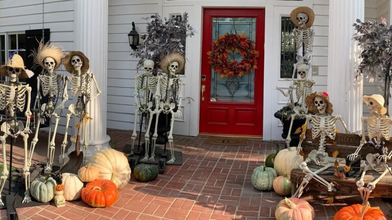 Skeleton figures in straw hats gathered on a white porch beside a red front door, decorated with a fall wreath and mixed pumpkins