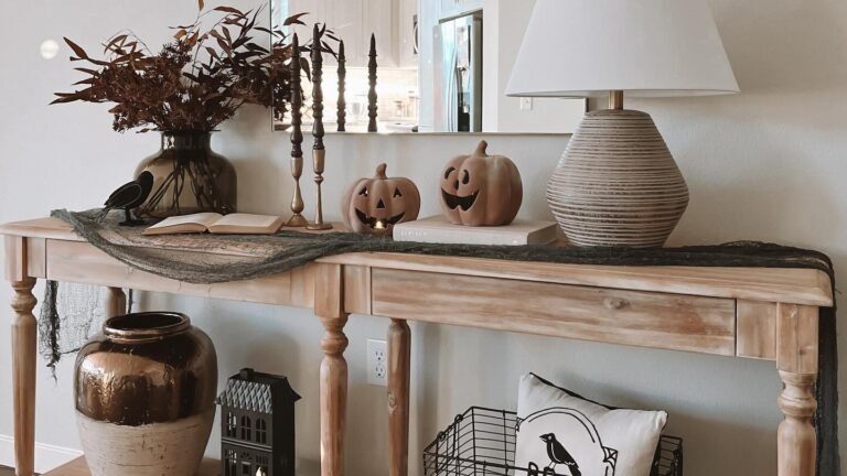 Terracotta jack-o-lantern pumpkins on a light wood console table with a dark cheesecloth runner, brass candlesticks, and a black bird figure for neutral Halloween decor