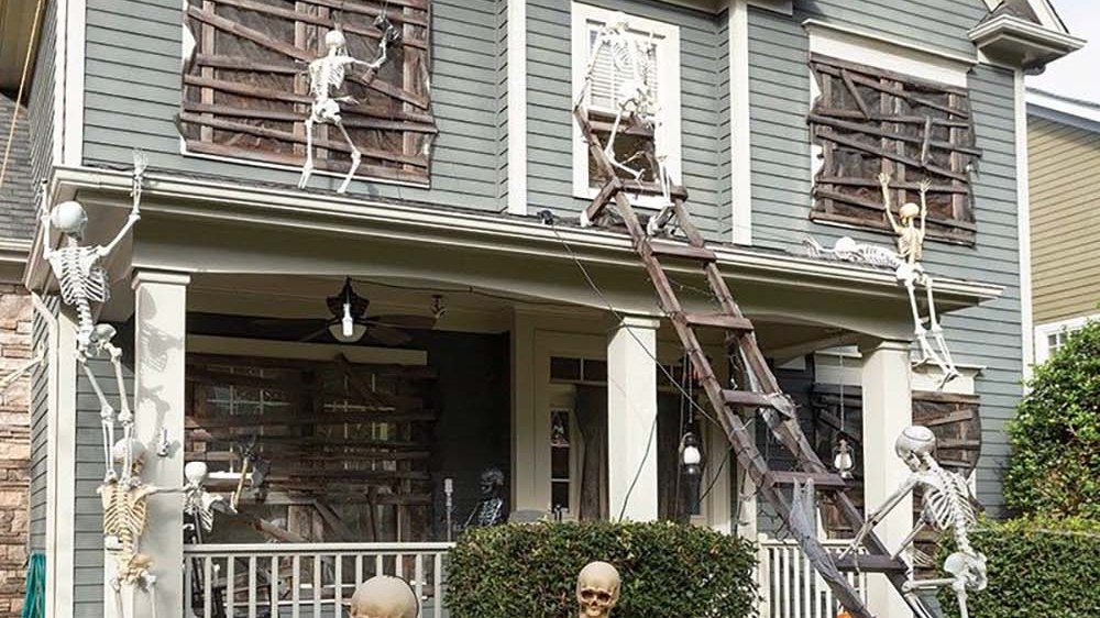 Skeletons breaking through boarded second-story windows and climbing a ladder on a two-story house, with a wheelbarrow of spare parts on the front lawn, full Halloween outdoor decor takeover