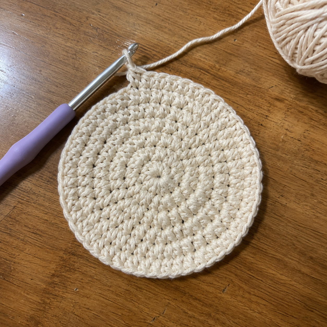 Flat crochet top disc lying flat on wooden table with purple crochet hook and cream cotton yarn