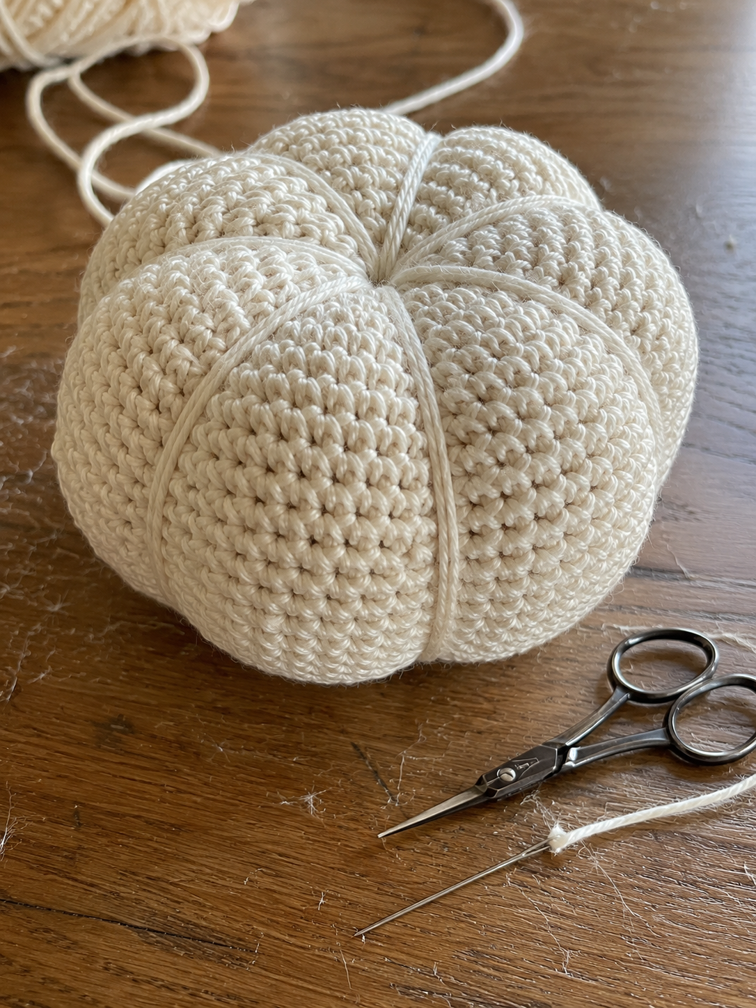 Close-up of cream crochet pumpkin showing six evenly-spaced ribs with scissors and yarn needle