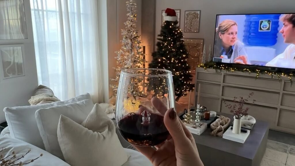 Holding a glass of red wine on a cozy couch with two small lit Christmas trees and a TV glowing in a warm apartment living room