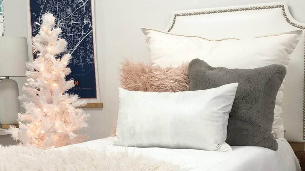 Small white Christmas tree glowing with warm white lights beside a twin dorm bed with a white duvet, a blush fur pillow, a gray fuzzy pillow, and a long white pillow in front of a studded headboard.