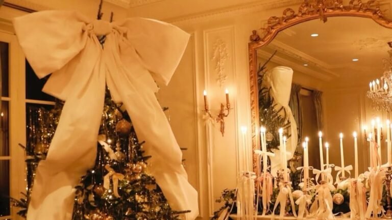 Oversized cream bow draped from the top of a lit Christmas tree with gold ornaments, beside a gilded antique mirror and a carved white mantel lined with taper candles tied with ribbons in a warm candlelit room.