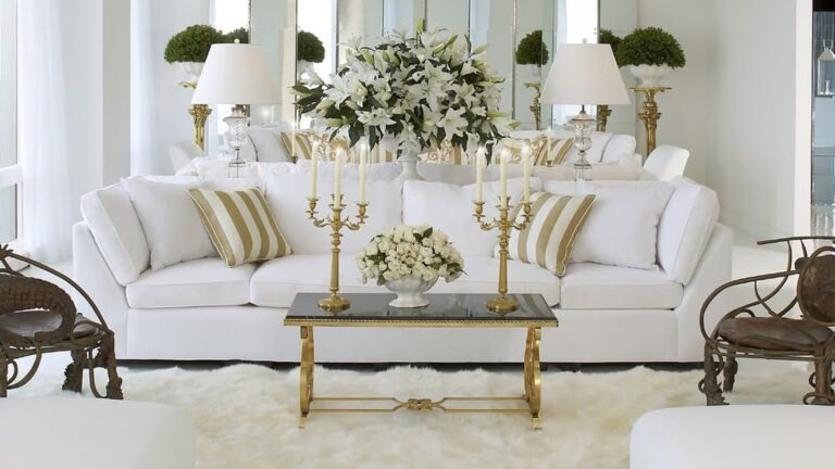 White sofa with gold striped pillows in a glam living room, styled with two gold candelabras and a gold coffee table, a big white lily bouquet behind it and a fluffy white sheepskin rug underneath.