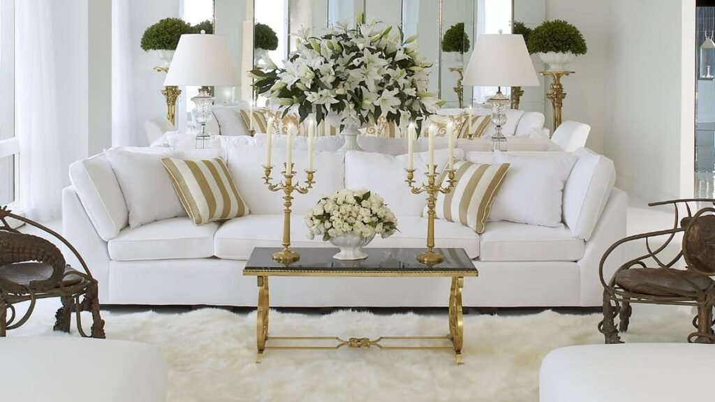 White living room with a plush white sofa, gold and cream striped pillows, a gold coffee table holding two gold candelabras and a bowl of white roses, a large white lily bouquet behind, and a fluffy white sheepskin rug underfoot.