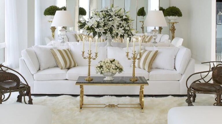 White living room with a plush white sofa, gold and cream striped pillows, a gold coffee table holding two gold candelabras and a bowl of white roses, a large white lily bouquet behind, and a fluffy white sheepskin rug underfoot.