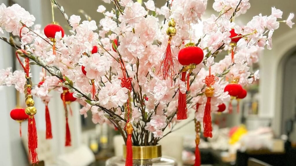 Tall vase of pink cherry blossom branches hung with small red velvet lanterns, red tassels and gold charms, set on a dining table in a bright home.