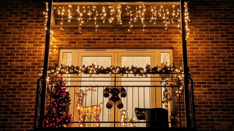 Brick apartment balcony decorated with Christmas lights at night, including white icicle lights hanging from the slab above, a pine garland with warm lights along the railing, a glowing gold reindeer and a small red-lit tree.