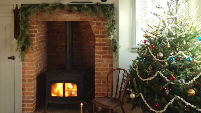Rustic Christmas room with an exposed red brick fireplace framed by a raw wood beam and pine garland, a black wood stove glowing with fire, and a fresh tree strung with popcorn garland and colorful glass ornaments.