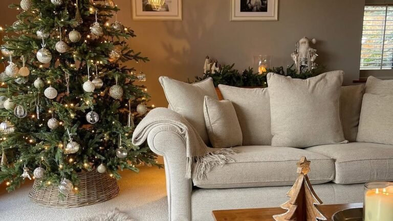 Christmas tree with silver and gold ornaments and warm white lights in the corner of a living room, next to a cream sofa with beige pillows and a draped throw, with a small wooden tree decoration on the table in front.
