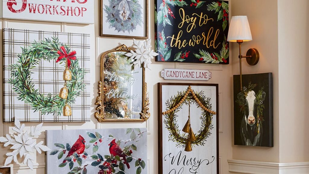 Christmas gallery wall of wreath paintings, a gold-framed mirror, a black Joy to the World canvas, red cardinal art, and a brass wall sconce, all hung on a cream paneled wall.