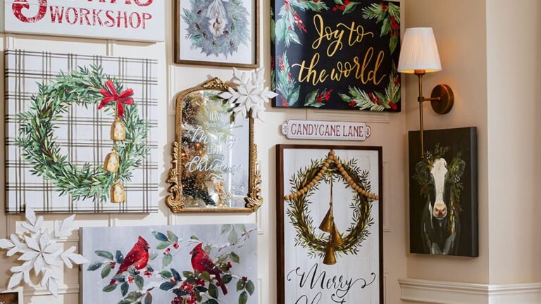 Christmas gallery wall of wreath paintings, a gold-framed mirror, a black Joy to the World canvas, red cardinal art, and a brass wall sconce, all hung on a cream paneled wall.