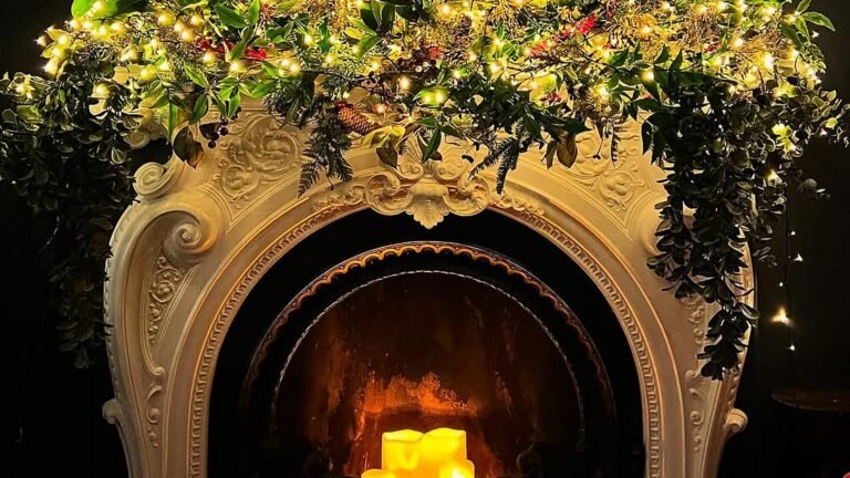 Ornate white carved fireplace surround with an arched opening, draped in a lit Christmas garland of green leaves, red berries and warm string lights, with glowing candles inside the firebox against a dark wall.