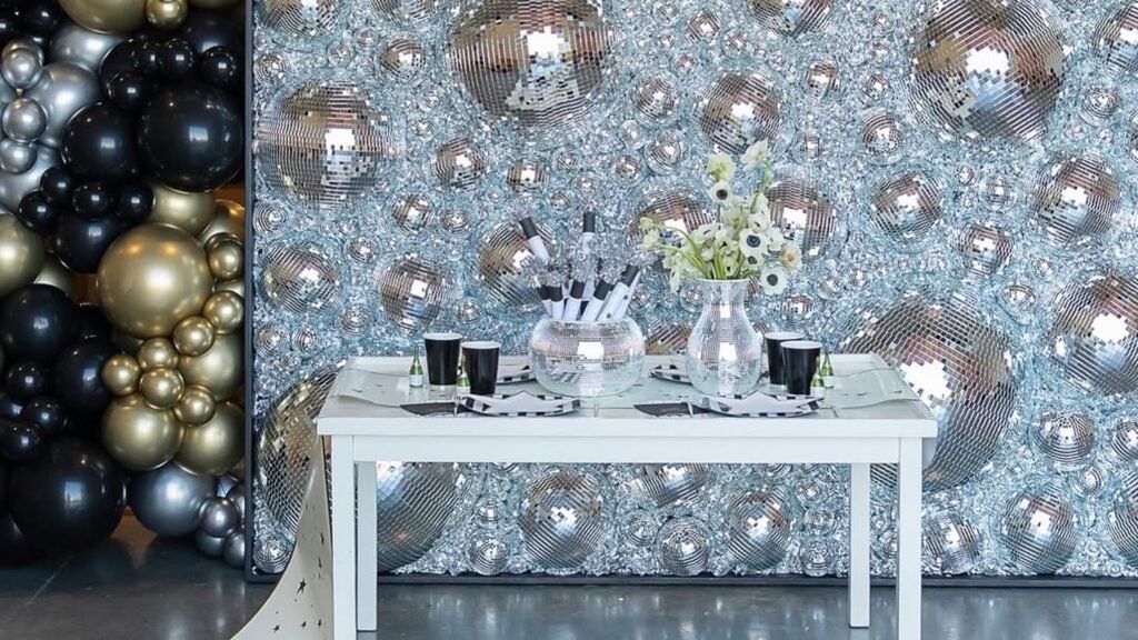 New Year’s Eve party backdrop with a wall of silver mirrored disco balls beside a black, gold and silver balloon garland, and a small white table in front holding black cups, a mirrored bowl of noisemakers and white flowers in a mirrored vase.