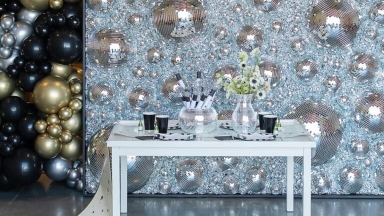 New Year’s Eve party backdrop with a wall of silver mirrored disco balls beside a black, gold and silver balloon garland, and a small white table in front holding black cups, a mirrored bowl of noisemakers and white flowers in a mirrored vase.