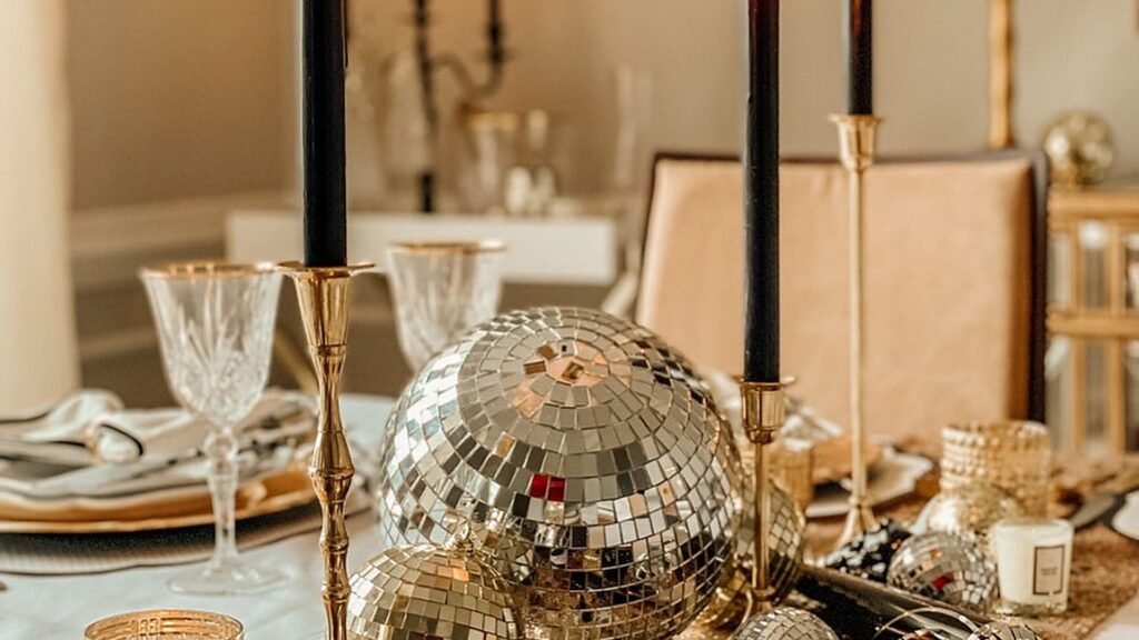New Year’s Eve dining table centerpiece with large mirrored disco balls, tall black taper candles in brass candlesticks, crystal wine glasses, and a gold glitter runner.