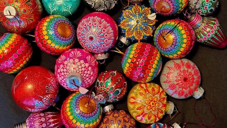 Pile of DIY Christmas ornaments hand painted with rainbow dots, pink and gold mandala designs, and red glitter, packed together on a dark fabric background.