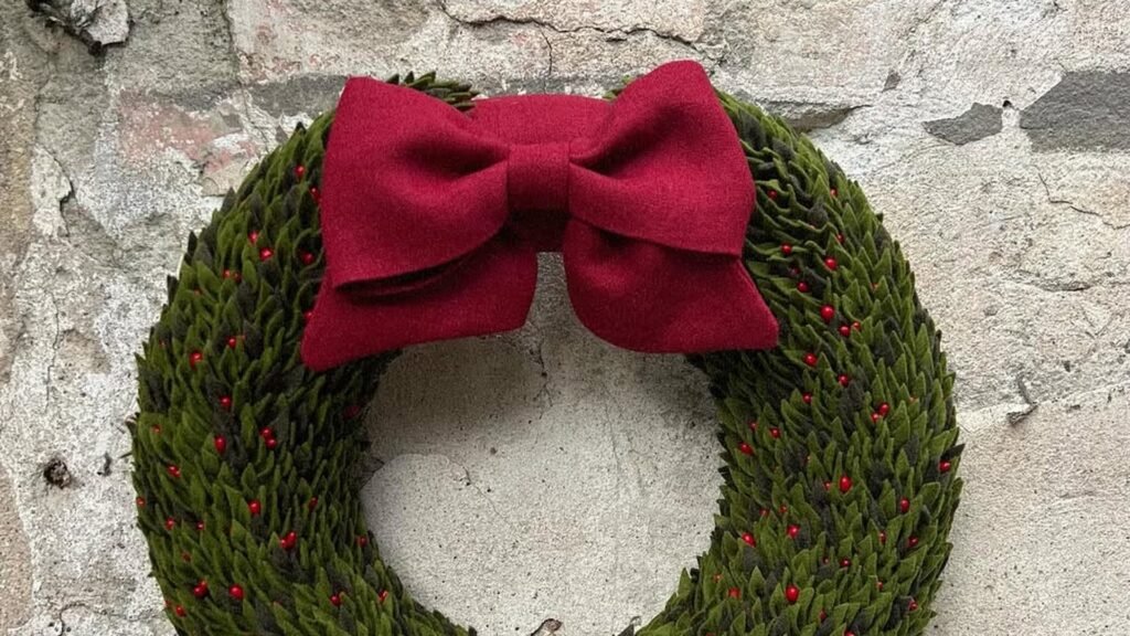 Round Christmas wreath made of overlapping olive green felt leaves dotted with tiny red berries, topped with a large burgundy bow and hanging on a rough gray stone wall.