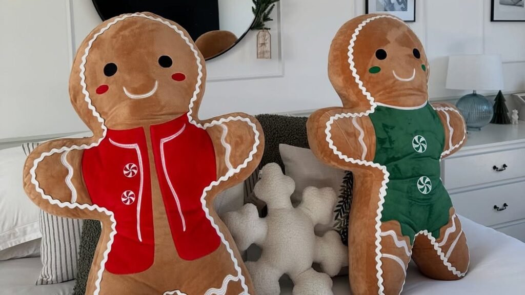 Two giant plush gingerbread men with smiling faces and white zigzag trim, one in a red vest and one in a green vest with peppermint buttons, sitting on a white bed beside a fluffy white snowflake pillow.