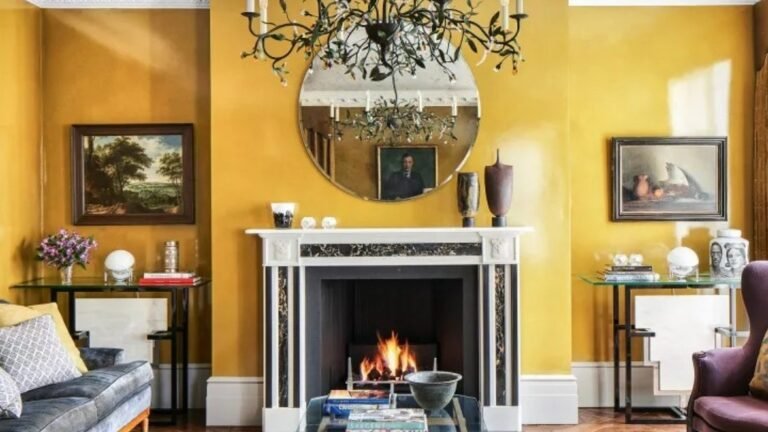 Glossy mustard yellow living room with a white marble fireplace and lit fire, a round mirror under a branch-style chandelier, framed oil paintings on both sides, and a blue-gray velvet sofa at the edge of the frame.