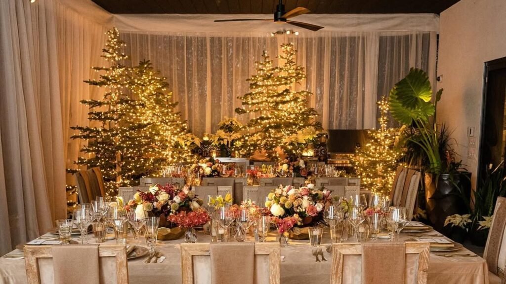 Warm-lit Christmas trees glowing behind a long dining table with a champagne velvet tablecloth, cream chairs with fringed throws over the backs, and low pink and cream flower centerpieces.