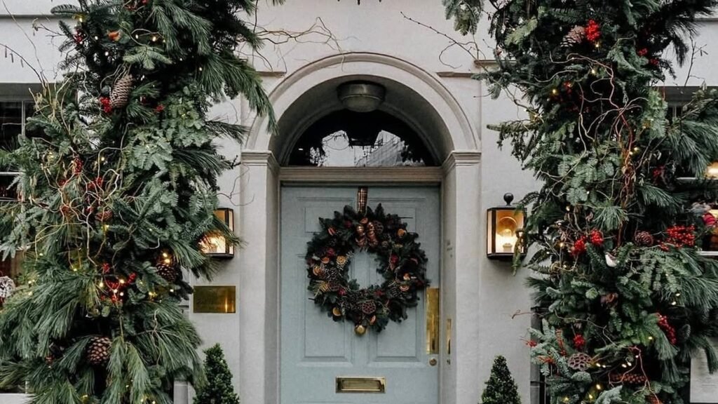 Light blue front door in an arched doorway with a round evergreen wreath of pinecones and dried orange slices, framed by thick pine and fir garlands with red berries, pinecones, and tiny white lights, and glowing lanterns on both sides.