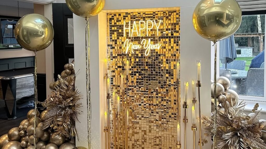 Gold sequin shimmer wall with a glowing white Happy New Year neon sign, framed by tall gold candle stands, two large gold orb balloons on long tassel tails, and gold balloon clusters with metallic leaves on a herringbone floor.