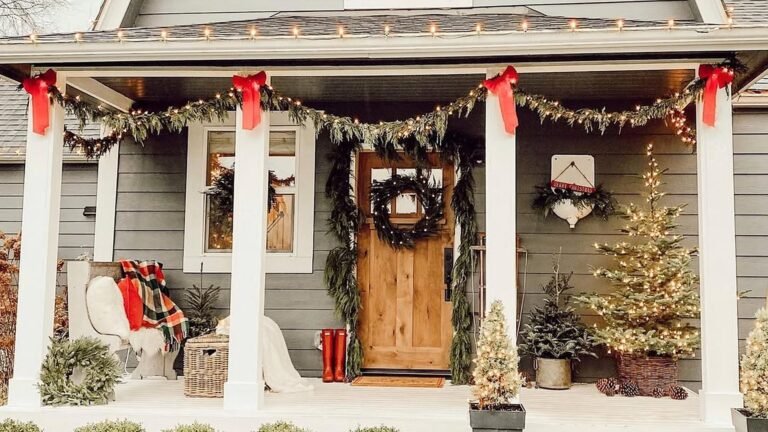 Christmas front porch with white columns tied with red bows, a lit evergreen garland swag, a black wreath on a natural wood door, and a glowing tree in a basket planter beside it.