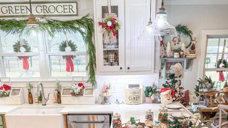 White farmhouse kitchen decorated for Christmas with green garland draped across and down the sink window, small wreaths with red bows in each window pane, and a striped ribbon wreath hanging on the glass cabinet door.