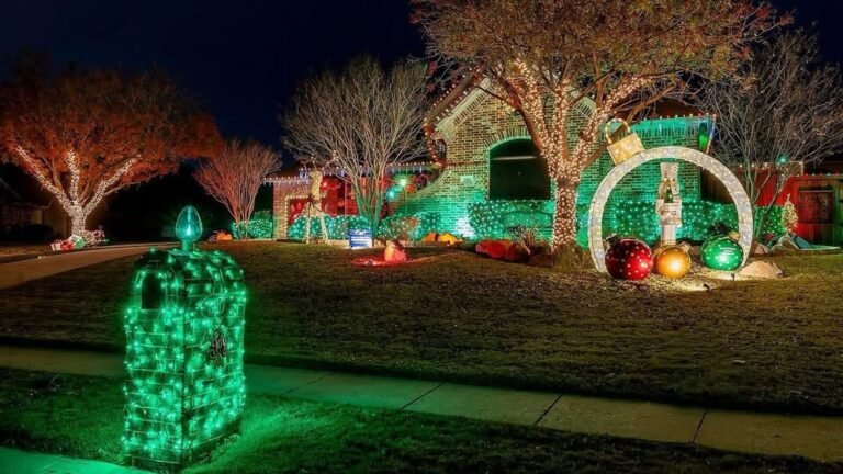 Mailbox wrapped in glowing green Christmas lights stands on a front lawn at night, with a brick house lit in green and red, trees wrapped in warm white lights, and giant ornaments under a lit ring.