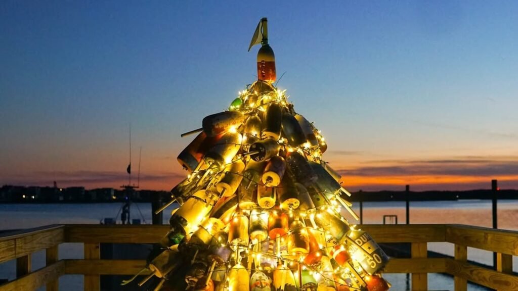 Christmas tree built from stacked colorful lobster buoys and wrapped in warm white string lights, glowing on a wooden pier against a deep blue and orange sunset over the water.