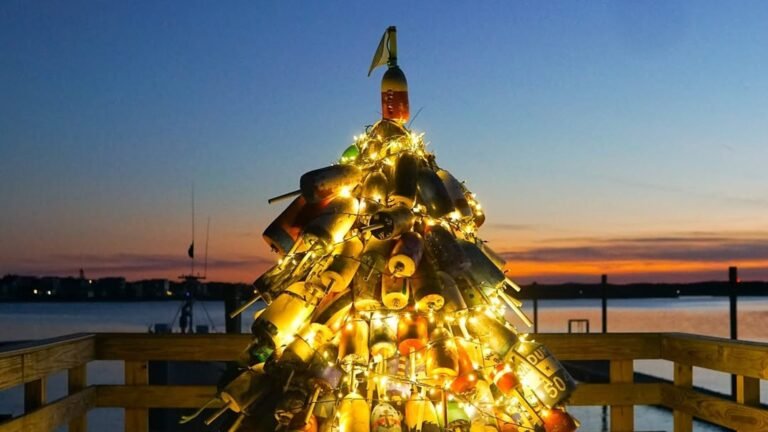 Christmas tree built from stacked colorful lobster buoys and wrapped in warm white string lights, glowing on a wooden pier against a deep blue and orange sunset over the water.