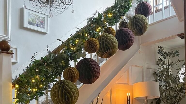 Olive green, mustard and plum honeycomb paper balls hanging from a white Christmas staircase railing beneath a leafy garland with warm fairy lights, with a glowing table lamp below.