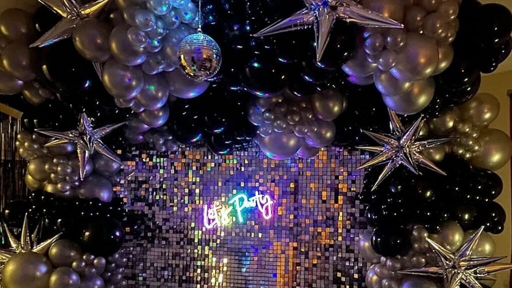 Black and silver balloon arch with silver star foil balloons and a hanging disco ball framing a shimmering sequin wall with a glowing neon Let’s Party sign for a New Year’s Eve party.