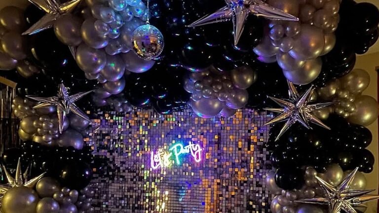 Black and silver balloon arch with silver star foil balloons and a hanging disco ball framing a shimmering sequin wall with a glowing neon Let’s Party sign for a New Year’s Eve party.