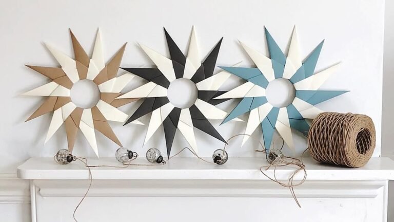 Three handmade origami paper stars in kraft brown, black, and slate blue with cream points leaning against a white wall on a white mantel, beside a string of small glass baubles on twine and a spool of brown paper cord.