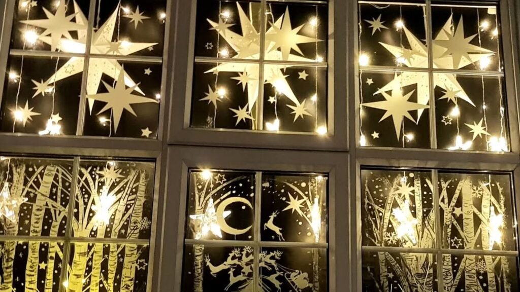 Christmas window decor with large white paper stars hanging in the top panes and a hand-drawn birch forest, crescent moon, reindeer and Santa sleigh on the lower panes, all glowing under warm string lights.